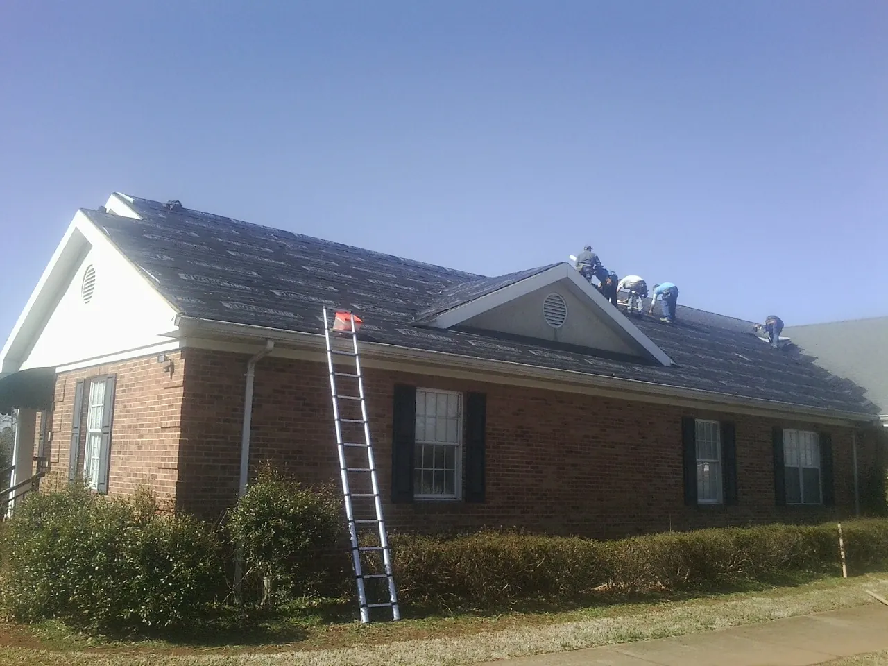 Crew tearing shingles off a brick ranch roof with a ladder set — Metal Roof Repair work by our crew in Treynor
