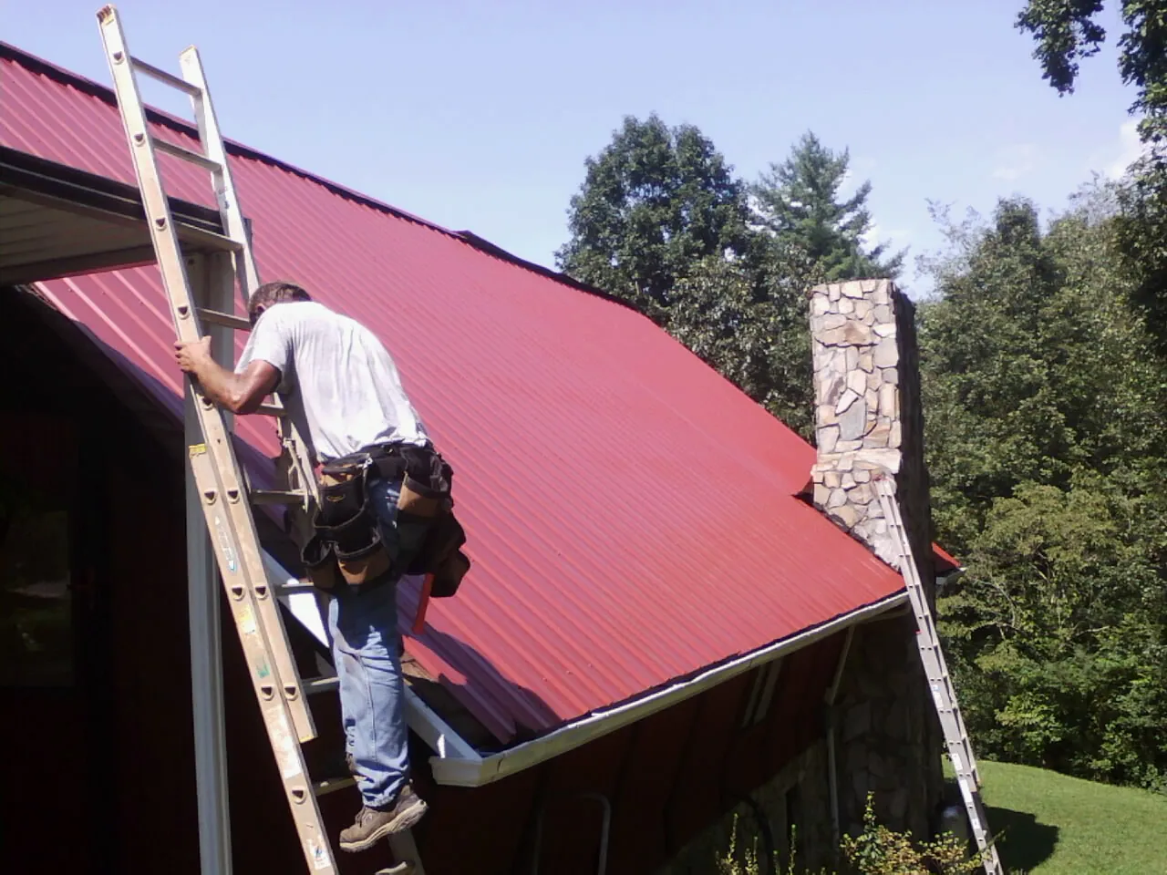 Roofer on a ladder beside a red metal roof and stone chimney — Soffit & Fascia Repair by Roofing Brothers in Treynor